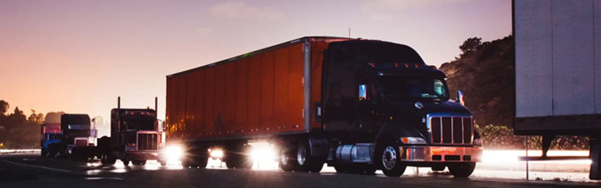 Truck sitting in a truck stop as the sun goes down in the distance. A chain of large big rig trucks one after another.