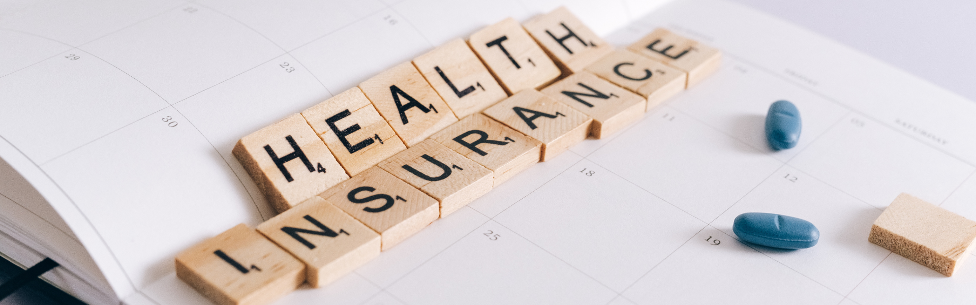 scrabble pieces that spell out "health insurance" sitting on a paper with a couple of pills