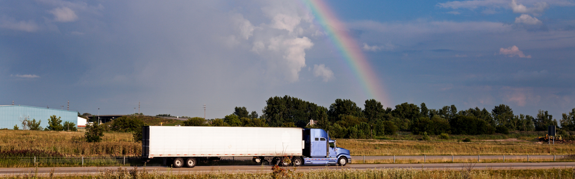 blue semi truck with white trailer driving on road with rainbow in background