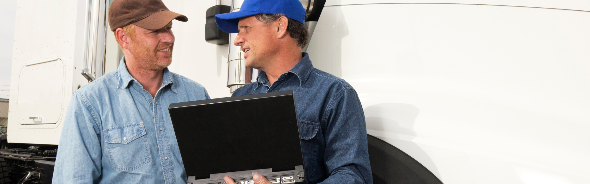two men in hats looking at laptop with white semi truck in background