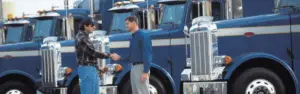 two men shaking hands with several blue semi trucks parked in the background