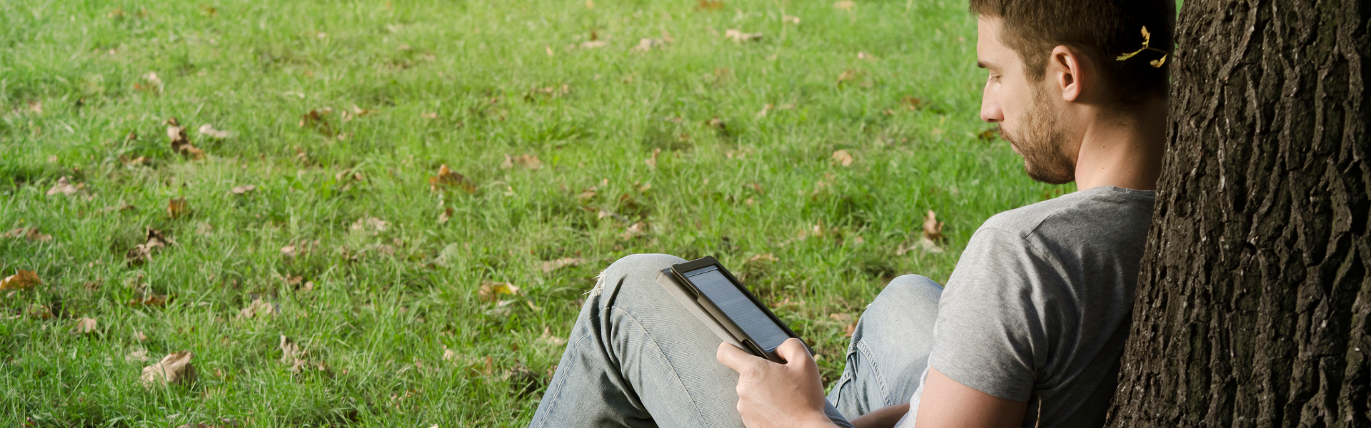 man leaning against tree reading ebook