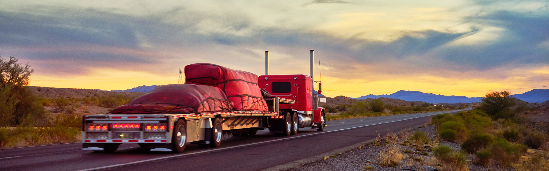 red flatbed truck with red cargo cover driving into sunset with mountains in the background