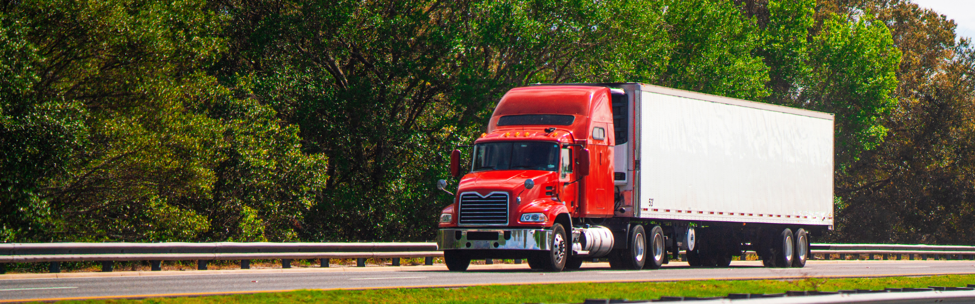 red semi truck pulling a white trailer on road with trees in the background