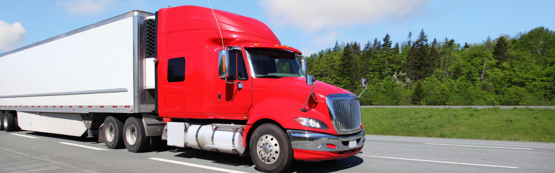 red semi truck pulling white trailer on road with green grass and trees in background