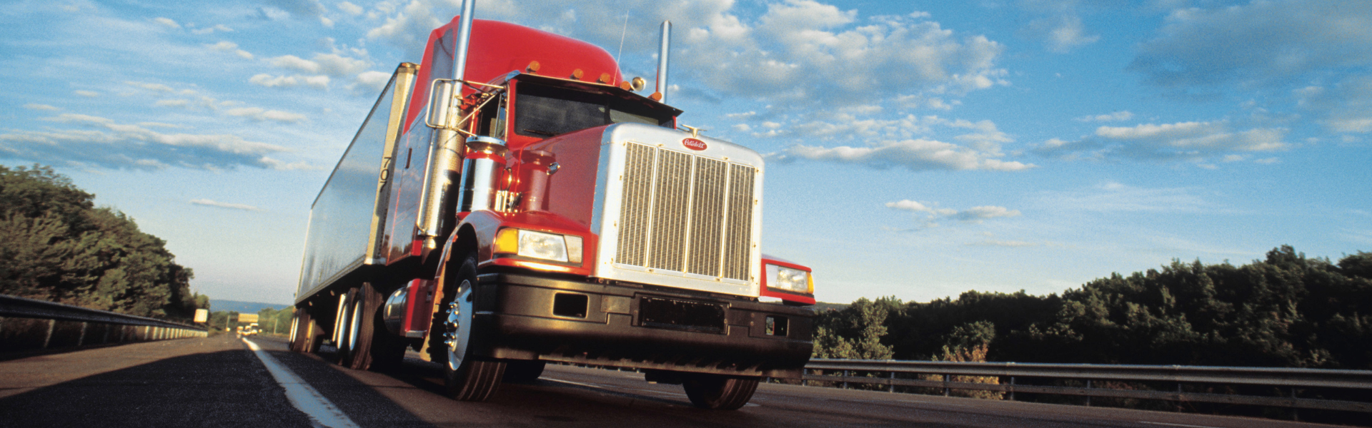 red semi truck on road trees in background and a blue sky with a few clouds