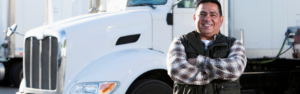 man in flannel shirt standing with crossed arms in front of white semi truck