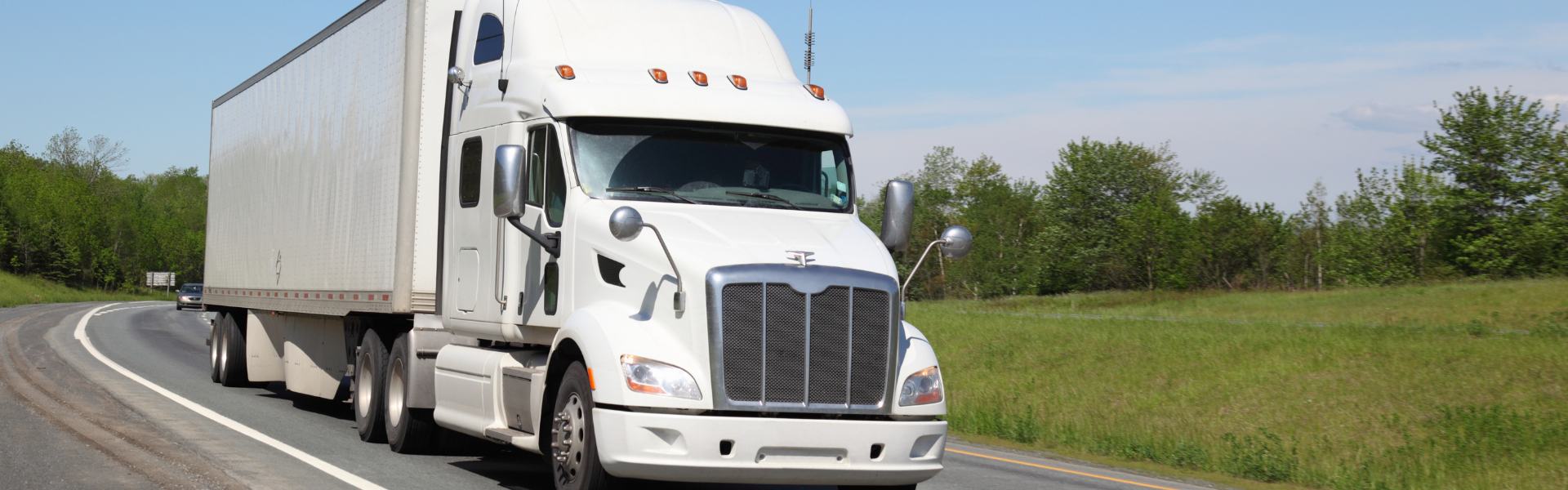 white semi truck with white trailer on curved road with green grass and trees in the background