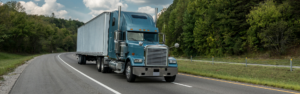 blue semi truck with white trailer driving on road with lush green background of trees and grass