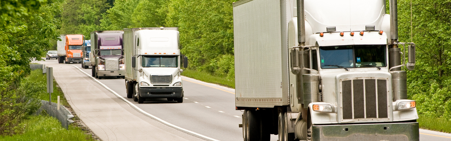 line of several semi trucks driving on highway with green trees in the background