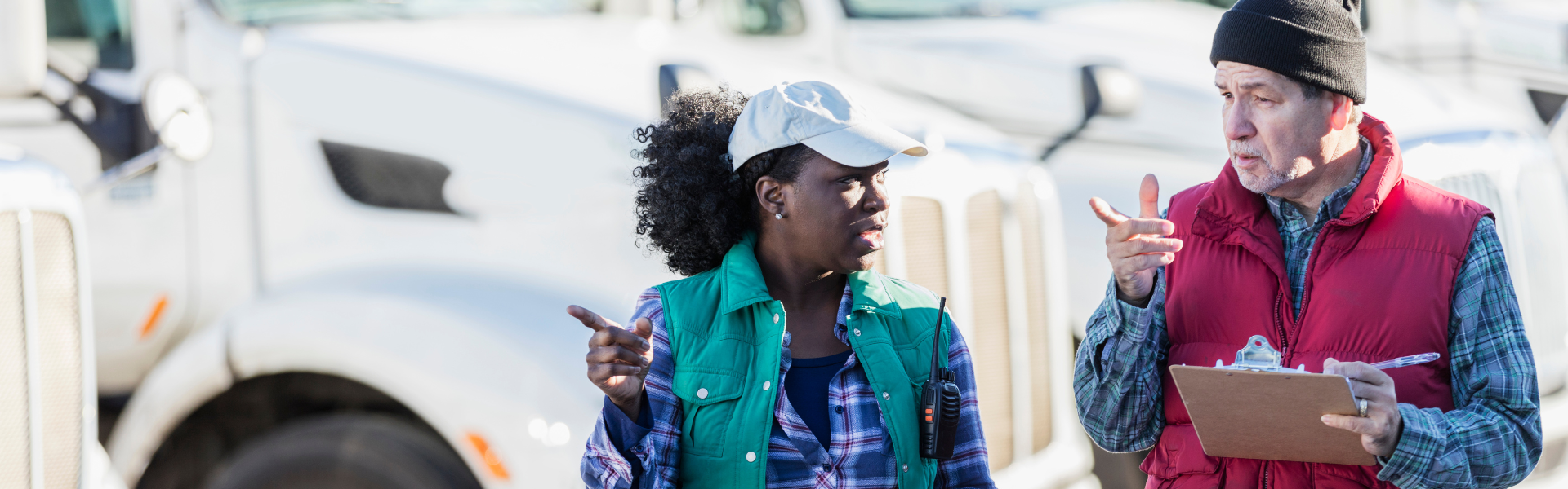 black woman wearing ball cap hat and flannel talking with man holding clipboard in front of parked semi trucks