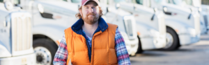 man in flannel shirt, orange vest, and hat standing in front of row of parked white semi trucks