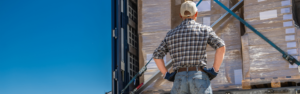 man in jeans, flannel shirt and ball cap hat standing with back to camera with hands on hips looking into loaded up semi truck trailer