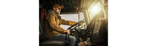 truck driver in cab of semi truck at the steering wheel driving with one hand on gearshift wearing coat, hat, jeans and sunglasses