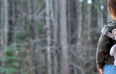 toddler with teddy bear backpack standing near edge of woods