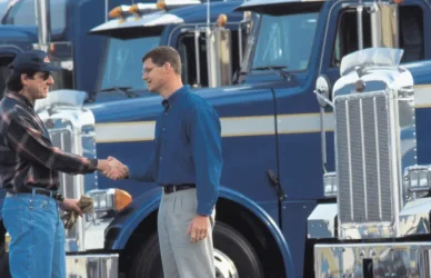 two men shaking hands with several blue semi trucks parked in the background