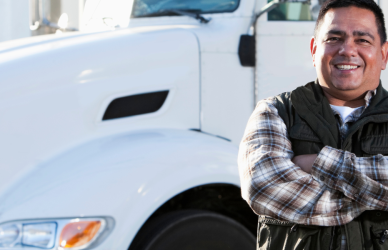man in flannel shirt standing with crossed arms in front of white semi truck