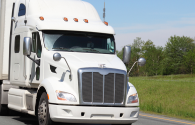 white semi truck with white trailer on curved road with green grass and trees in the background