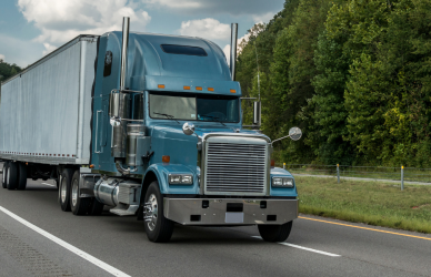 blue semi truck with white trailer driving on road with lush green background of trees and grass