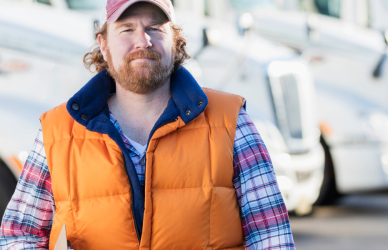 man in flannel shirt, orange vest, and hat standing in front of row of parked white semi trucks
