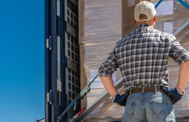 man in jeans, flannel shirt and ball cap hat standing with back to camera with hands on hips looking into loaded up semi truck trailer