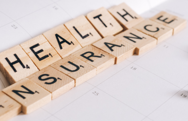 scrabble pieces that spell out "health insurance" sitting on a paper with a couple of pills