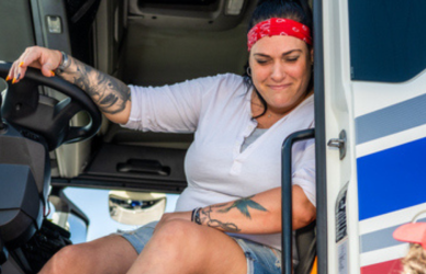 woman with tattoos sitting at the wheel in the cab of a semi truck with the door open
