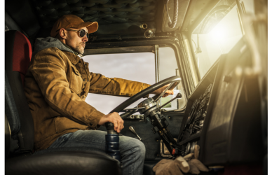 truck driver in cab of semi truck at the steering wheel driving with one hand on gearshift wearing coat, hat, jeans and sunglasses